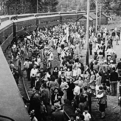 Black and white photograph. A passenger train at a station. A large crowd of stands alongside, some people talk to those already on board the train.