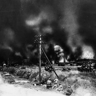 Black and white photograph. A road and open field. Smoke and many explosions are visible in the distance.