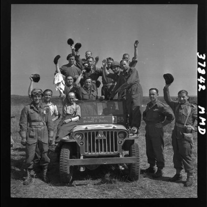 Black and white photograph. A group of men stand on and around a military jeep in the middle of an empty field. Most raise their caps in greeting to the camera.