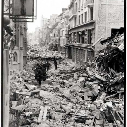 Black and white photograph. A line of Canadian troops proceeds down a narrow street in Caen. The buildings on either side have a lot of visible damage. The streets are completely covered with large pieces of rubble.
