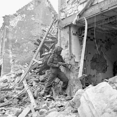 Black and white photograph. A soldier stands on a large pile of rubble pointing a gun into a gaping hole in the front of a building.  Another ruined building is visible behind him, and the street is entirely covered in rubble.