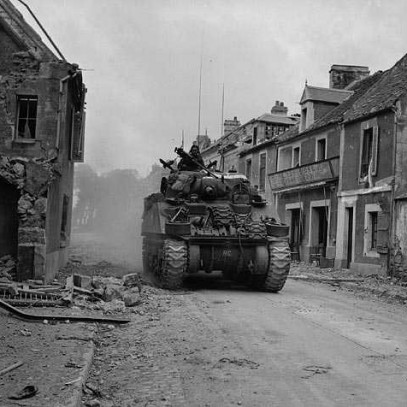 Black and white photograph. Buildings on a city street show significant damage. A Sherman tank navigates the rubbled-filled street.