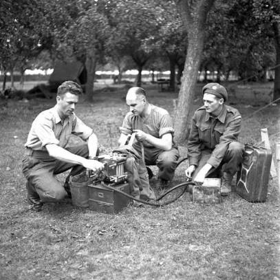 Black and white photograph. Three soldiers crouch on the ground, interacting with a small piece of machinery.