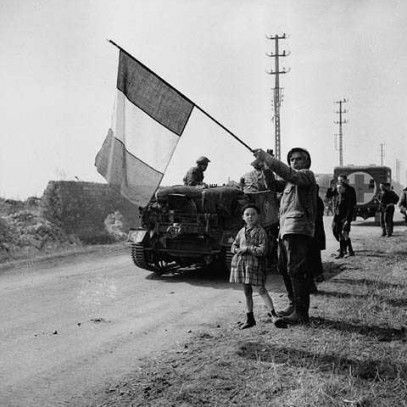 Black and white photograph. A French man stands with a young boy on the road side, holding a large French flag in the area. Soldiers file past on foot and in military vehicles.