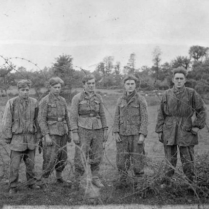 Black and white photograph. Five very young German soldiers in varying styles of battle dress stand behind a thickly laid barbed wire.