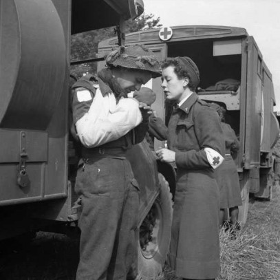 Black and white photograph. A nurse and a soldier stand in front of a military truck. The soldier’s right arm is in a sling, and both are examining it. Other trucks, nurses, and soldiers are visible in the background.