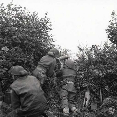 Black and white photograph. Three soldiers crouch in thick bushes. Two position a gun in a narrow gap. The third soldier looks off to the side. Equipment, weapons, and ammunition are on the ground.