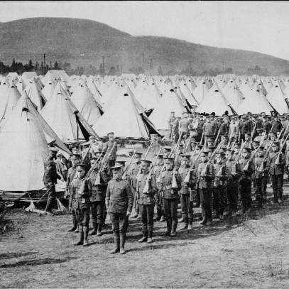 Black and white photograph. Soldiers stand at attention in an orderly column in front of a field of white cone shaped tents.