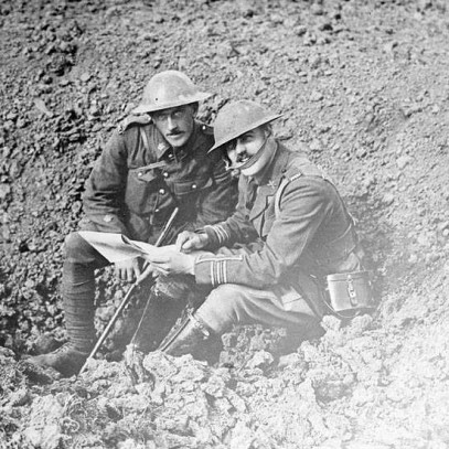 Black and white photograph. Two Canadian soldiers take a break from consulting the paper in their hands; they look at the camera from the shell-hole they are sitting in.