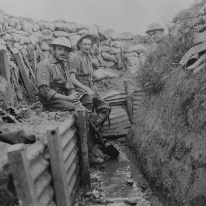 Black and white photograph. Two Canadian soldiers sit on a ledge in a muddy trench, while a third peaks from behind a corner. The floor of the trench is covered in water.