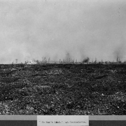 Black and white photograph. Shells can be seen exploding in the distance over barren No-mans land (labelled as such at the bottom of the image). The ground is muddy and uneven, dead trees are scattered.