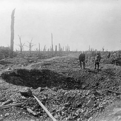 Black and white photograph. A large shell hole in the middle of the road. Two men in military uniforms and helmets stand behind it, one crouching slightly. The landscape is rough and rocky.