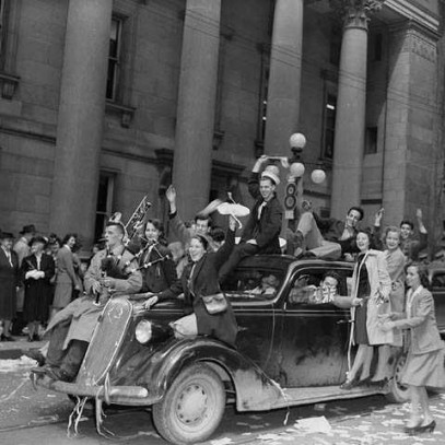 Black and white photograph. A car drives down the street over tickertape. Happy people ride inside and on top of it. Others crowd the street in front of a large concrete building.