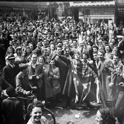 Black and white photograph. With storefronts in the background at an intersection, the streets are packed with people, several with bicycles, most facing toward the camera. Their faces are jubilant, and many wave flags. Hands are raised in celebration.