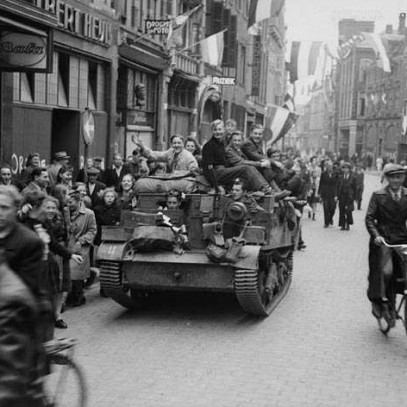 Black and white photograph. Street scene. Dutch flags are everywhere, in hands and hanging. People crowd the sidewalks and streets on foot and bike. A tank drives down the centre of the street, and civilians run alongside and hang off of it.