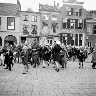 Black and white photograph. Cobblestoned town square, narrow building in background. A large group of children in warm coats circulate and move through the square, looking generally excited. Many wave small dutch flags. There is a Dutch flag hanging on a