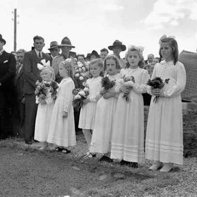 Black and white photograph. 6 young Dutch girls stand in a row, wearing white dresses and holding bouquets of flowers. Onlookers in civilian and military clothing stand behind them.