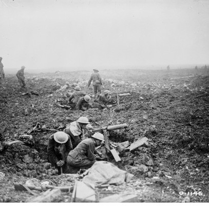 Black and white photograph. Two large holes in the ground. In each one, 3-4 men in military gear arm and aim a machine gun. The ground is muddy and the air is filled with smoke. Several men walk behind them.