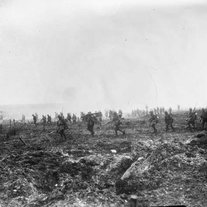 Black and white photograph. A large number of men walk, spaced out, through a misty day on very muddy and broken ground. Several carry large guns on their shoulders.