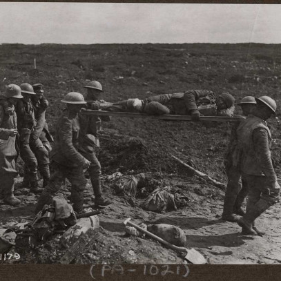 Black and white photograph. A muddy battlefield. Four men in military uniforms and helmets carry a man on a stretcher on their shoulders. Three men follow behind them, the man in the centre supported by the other two.  Debris litters the ground.