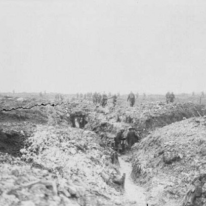 Black and white photograph. A photo of a large, muddy field with a Y shaped trench. Men, distant from the camera, walk through the trenches and look into dugouts. Beyond the trenches, men are visible in silhouette walking across the landscape.