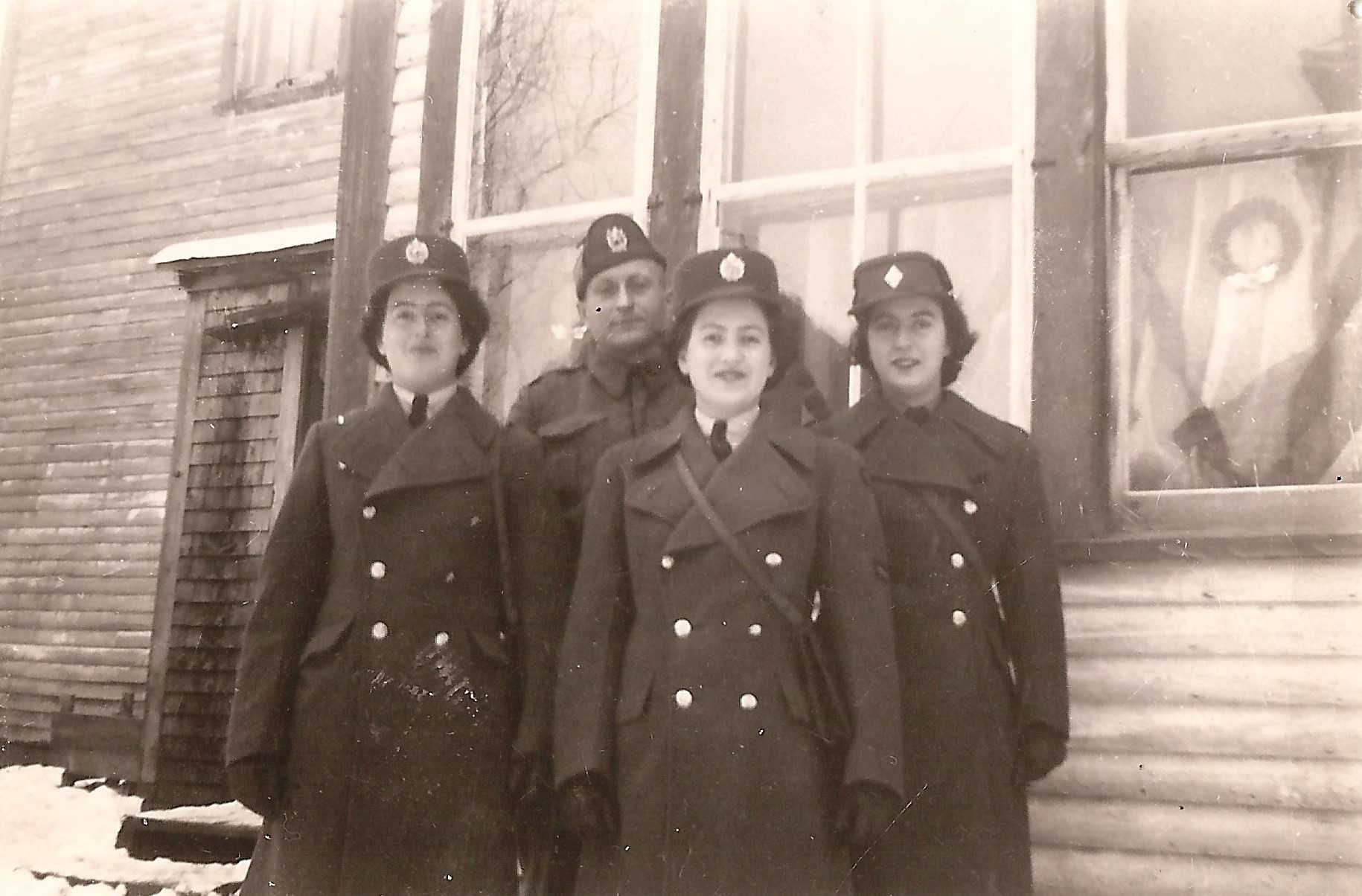 Black and white photograph. Three young women stand in front of their father, in front of a house. They are all in winter military uniforms and their is snow on the ground.
