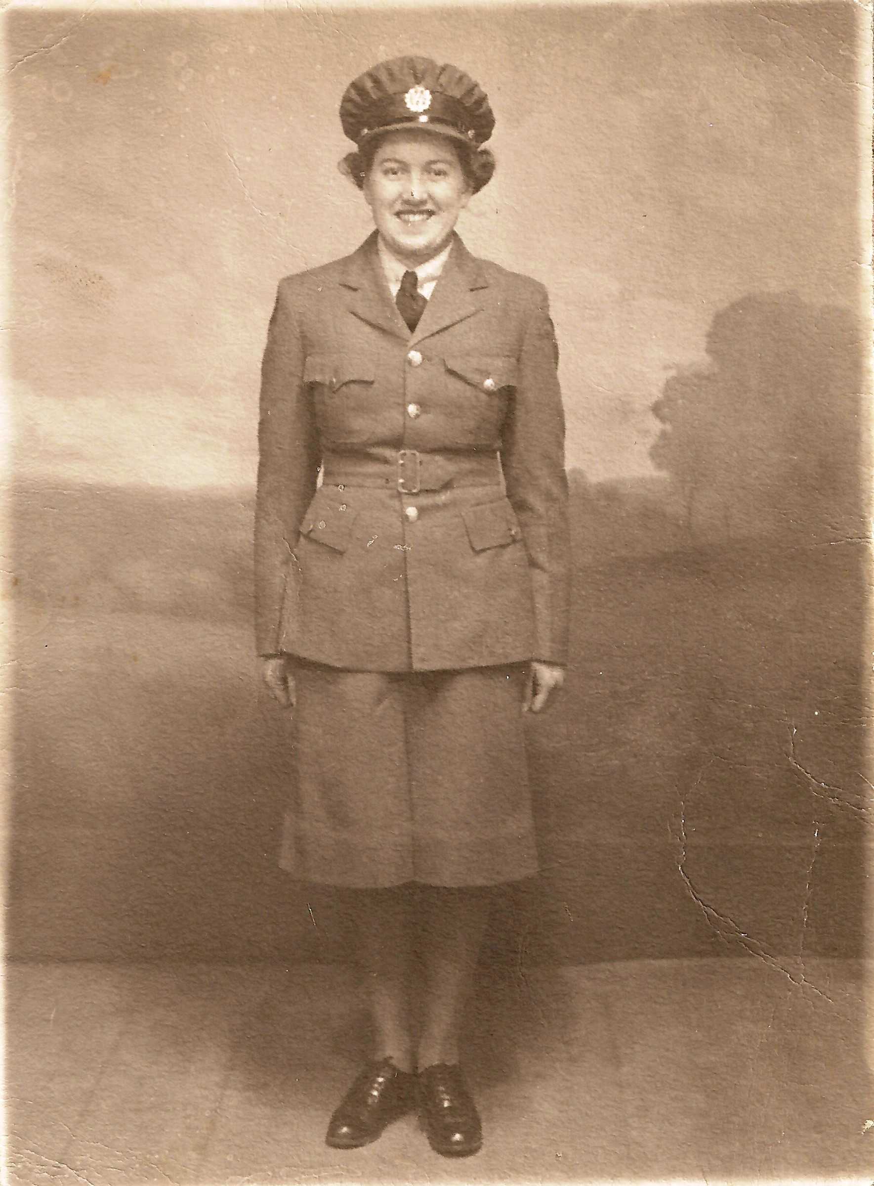 Sepia tone photo. A young women is shown in full military dress, in a skirt uniform. She is grinning standing at attention.