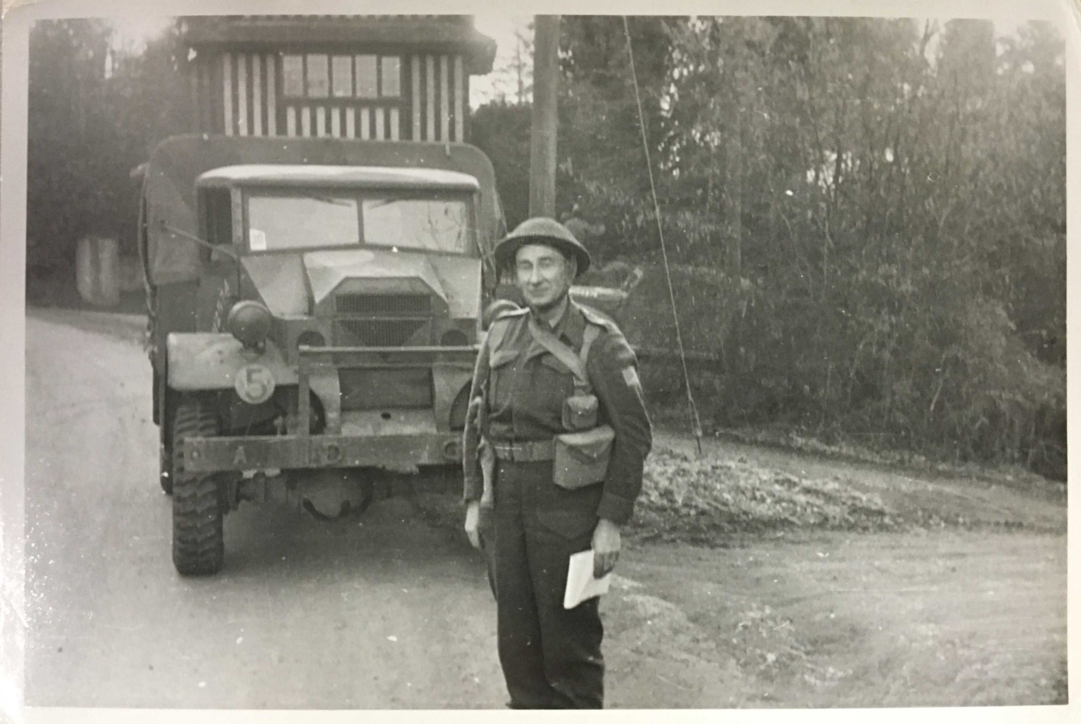 Black and white photograph. Archie stands at attention, with a smile, in front of a military vehicle.