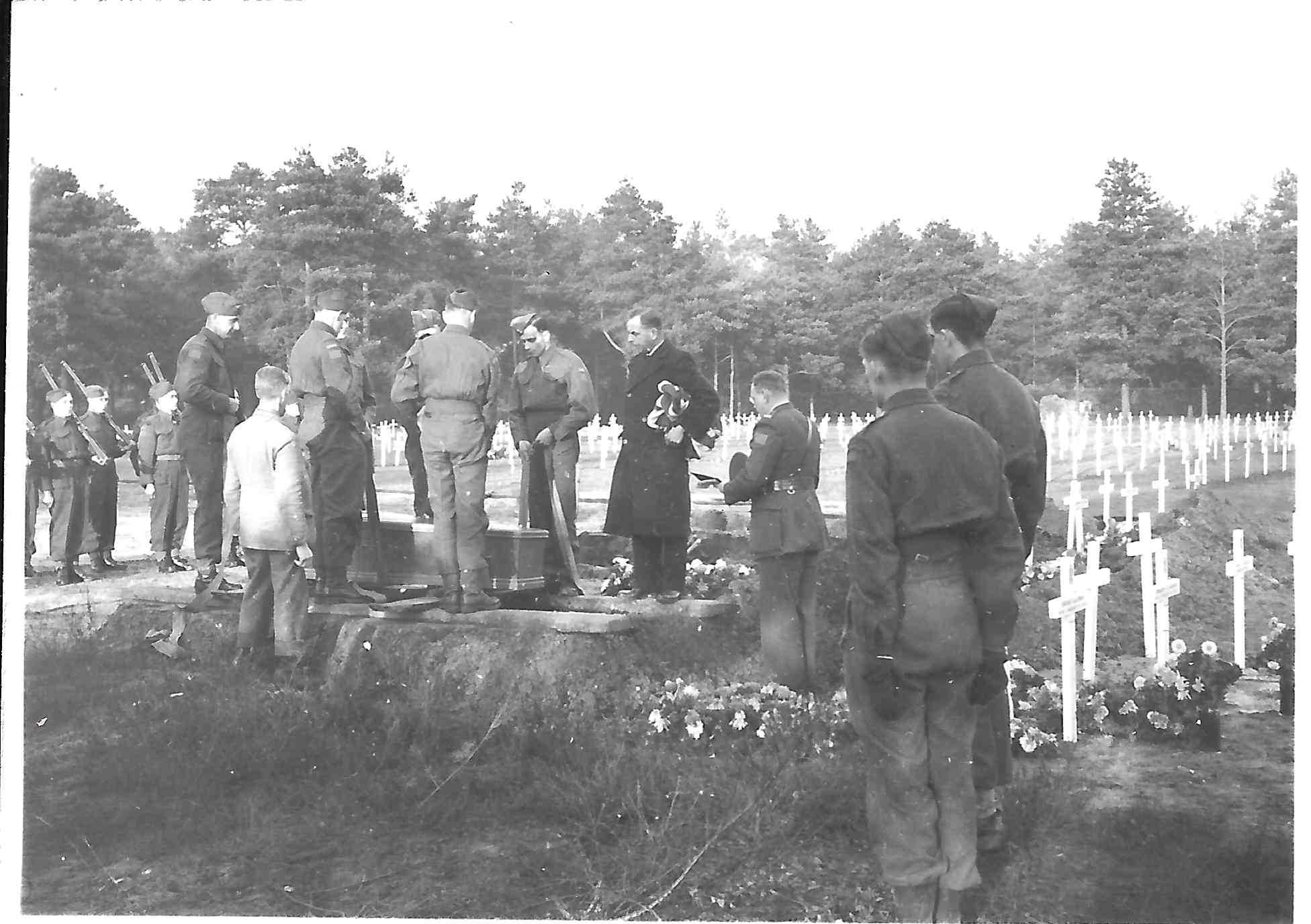 Black and white photograph. Men in various uniforms stand among a field of white crosses, lowering a coffin into a grave. A military padre stands at the head of the grave.