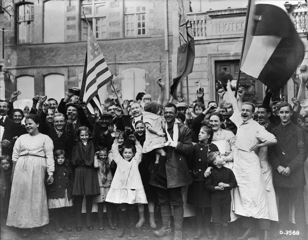 Black and white photograph. Canadians in Valenciennes amongst a crowd of cheering French citizens. They wave various allied flags. The faces of men, women, and children are all joyous.