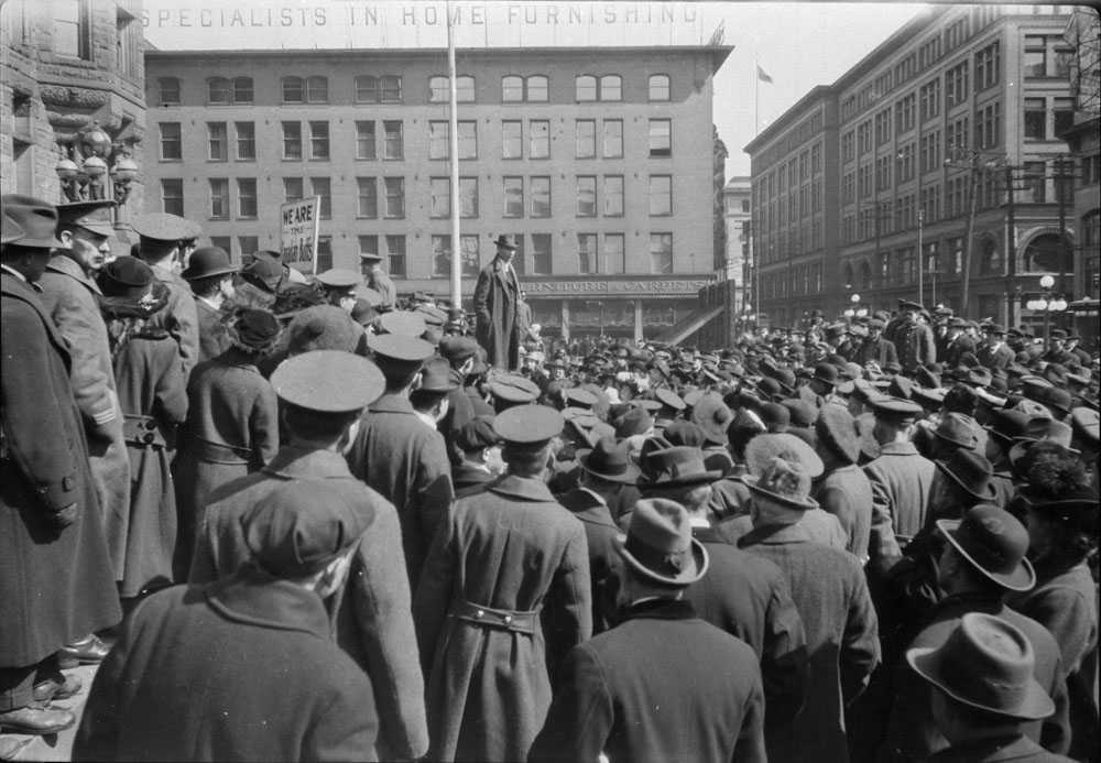 Black and white photograph. Thousands gather at a recruiting rally in downtown Toronto. Some are in uniform, others are in civilian clothing. One man stands higher than the rest in the centre of the photograph, making a speech.