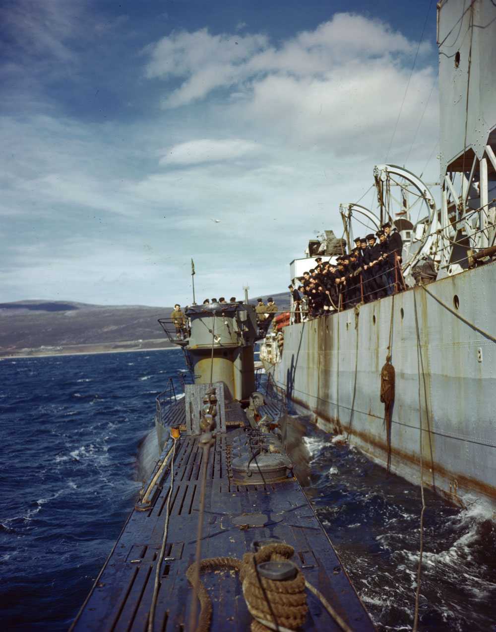 Colour image. The side of a large ship is shown, Canadian sailors lean over the rail on the side. Attached to the boat by ropes is a barely-surfaced submarine; German military personnel are standing on top of it.