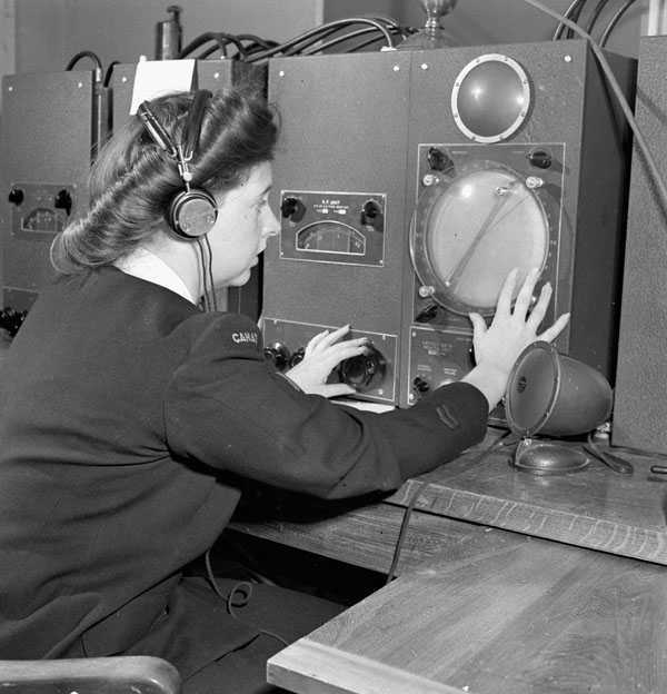 Black and white photograph. A woman with styled hair and manicured nails watches a readout on a piece of equipment, using one hand to turn a dial. She is sitting at a large wooden desk and surrounded by similar machines. She is in military dress.
