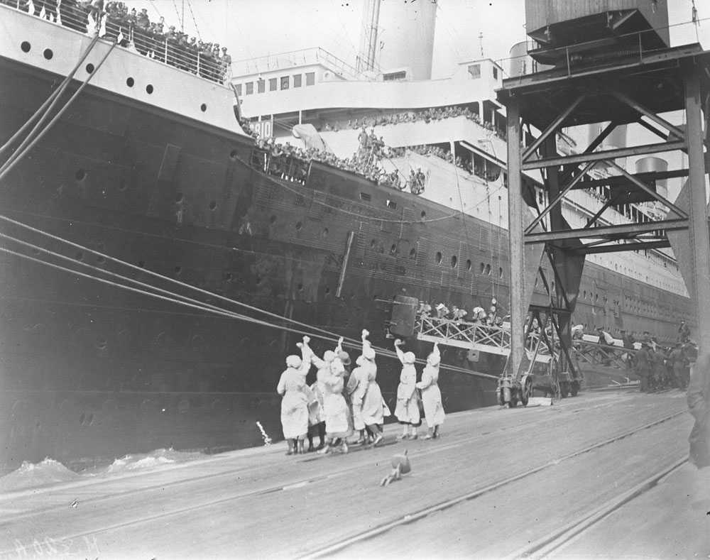 Black and white photograph. Women wave handkerchiefs on a dock while men board a ship.