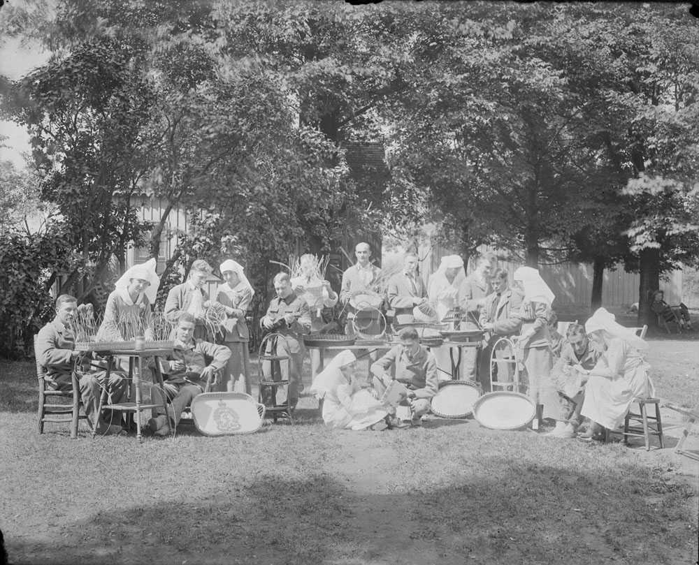 Black and white photograph. Nurses and soldiers sit on chairs or on the ground near a large tree. They ware weaving baskets.