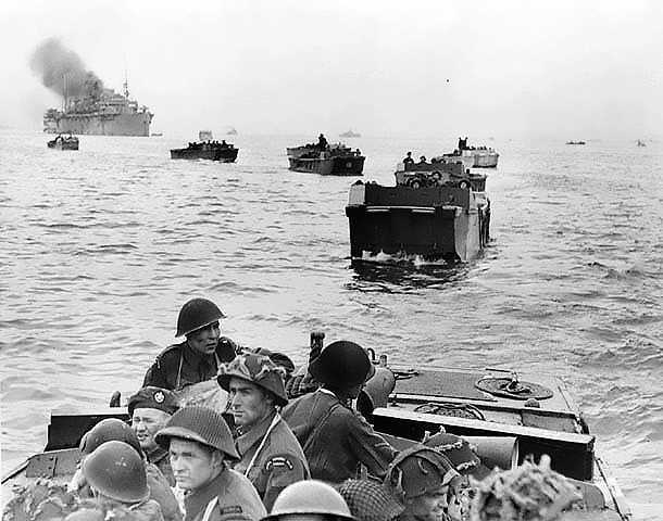 Black and white photograph. A row of landing craft float away from a large boat. Soldiers gaze out at the open sea.