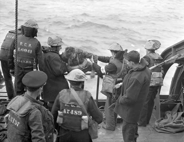 Black and white photograph. The edge of a boat, wavy water below. A soldier is pulled onto the boat by members of the Royal Canadian Navy, most wearing life jackets. Members of the Royal Canadian Medical Corps are identified by. their red cross arm bands.
