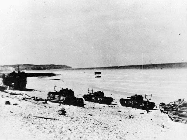 Black and white photograph. 4 tanks and a landing craft are abandoned on the rocky beaches of Dieppe. In the distance, military equipment sitting in the shallow tide is visible.