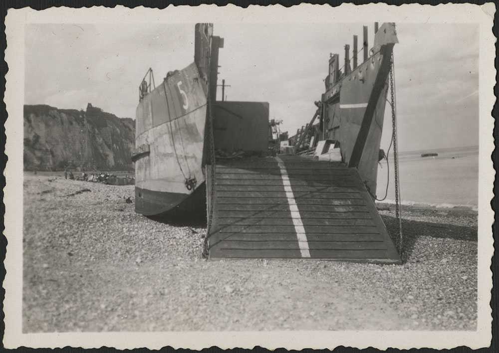 Black and white photograph. A landing craft on the pebbly beaches at Dieppe, landmark white cliffs in background. The craft is fully on the beach, and it's landing platform is down.