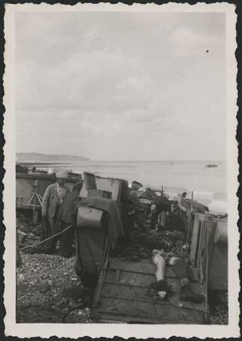 Black and white photograph. Civilians of Dieppe are shown loading dead Canadian soldiers into a vehicle on the beach. The water is visible in the background.