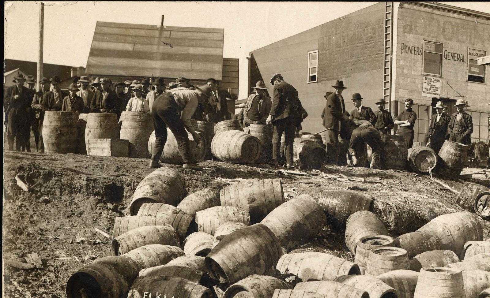 Black and white photograph. A pile of empty barrels sit at the shore of a lake. Slightly up hill, men are crowded around more barrels, moving them towards the lake for emptying.