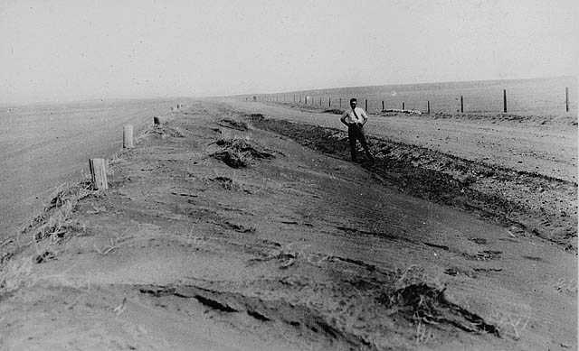 Black and white photograph. A man stands on the side of a road, flanked by fields. Despite the black and white colouring of the photo, it is easy to tell how dry the ground is. Dirt drifts through the air.