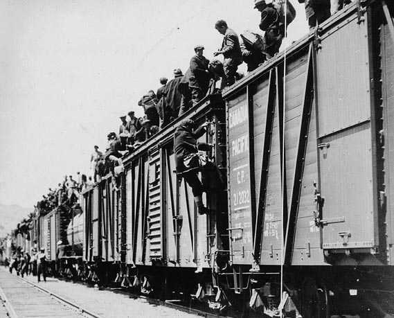 Black and white photograph. Cargo train cars on a track, groups of men mingle nearby. Hundreds of men sit on top of the cars.