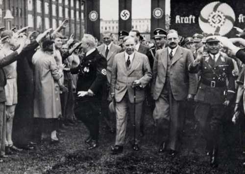 Black and white photograph. Prime Minister King walks through a crowd of civilians flanked by Nazi officials.