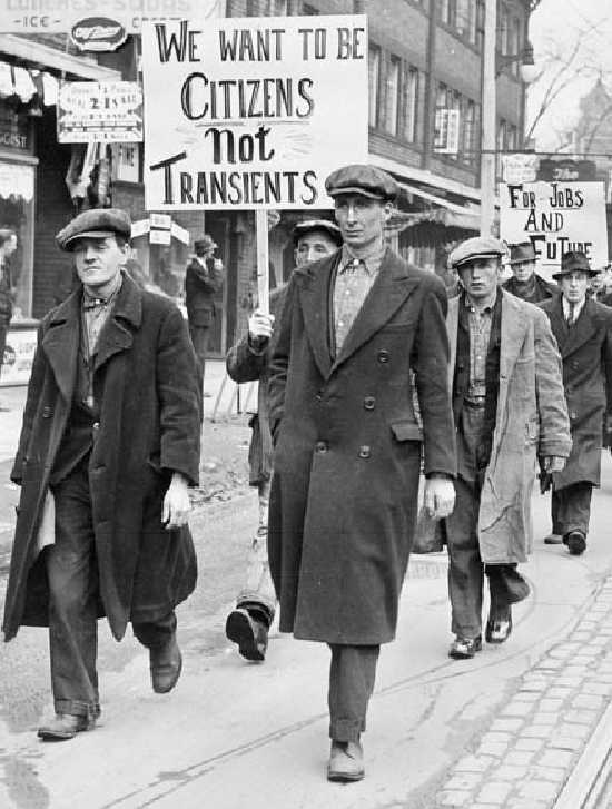 Black and white photograph. Men in long coats march down a city street. Several hold signs.