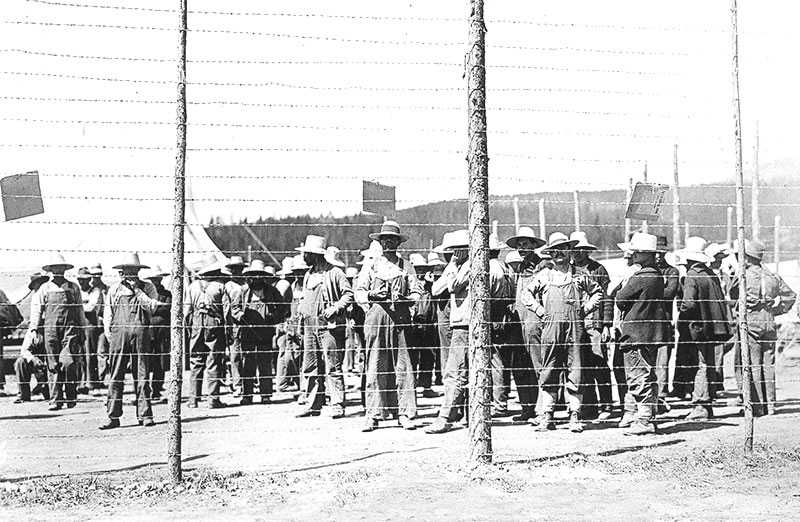 Black and white photograph. A large group of men, dressed in overalls and wide-brimmed hats, stand idly behind a high barbed wire fence. A tent is visible in the background.