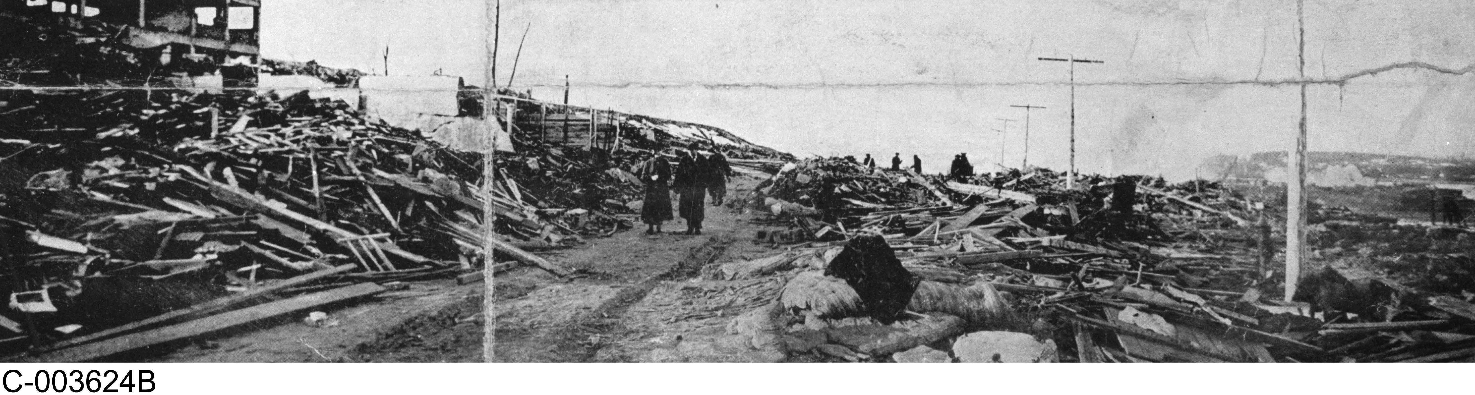 Black and white photograph. Survivors walk along a narrow path through the debris, away from the shoreline. The skeletal remains of a large building are on the left.