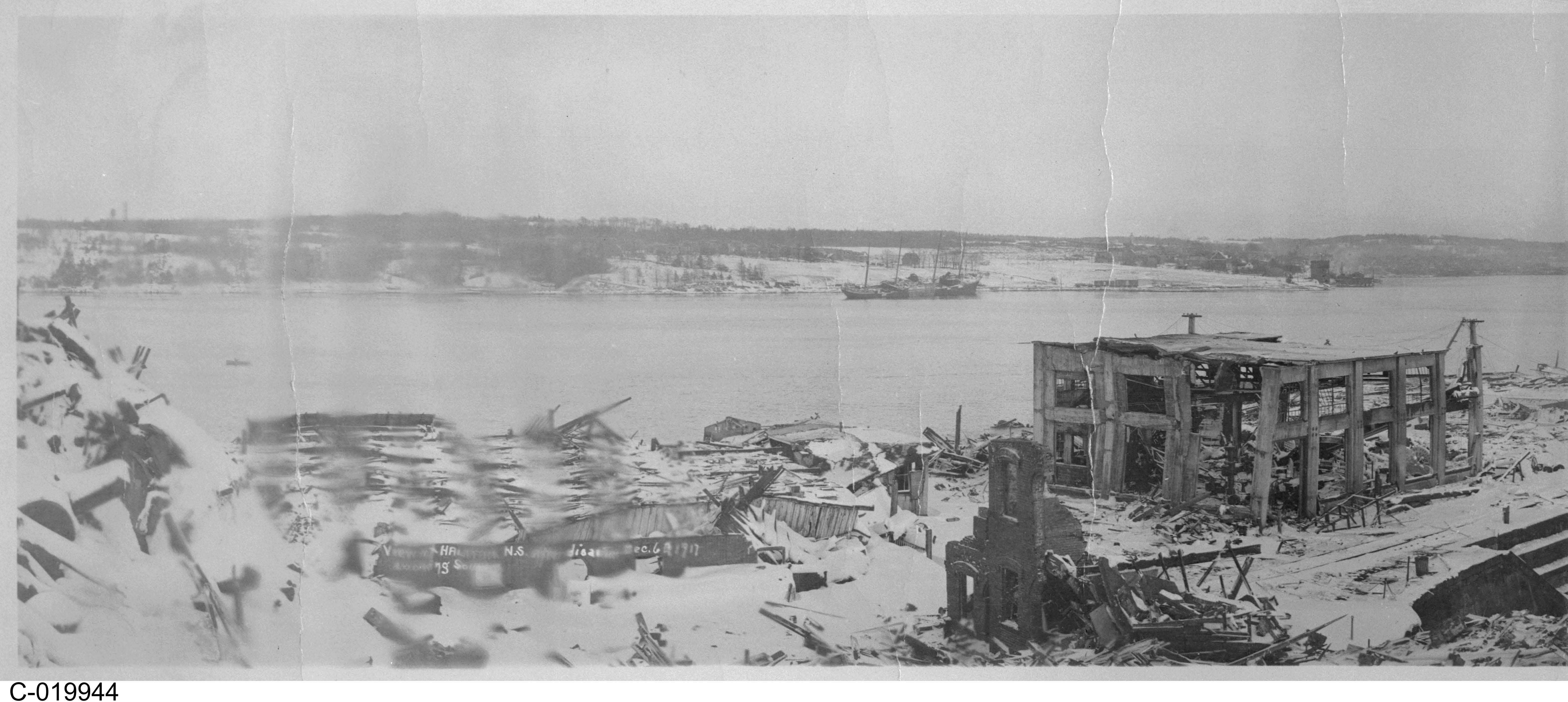 Black and white photograph. A variety of buildings are shown in ruins. Factories are in operation in the distance. A body of water is visible on the left side of the photo.