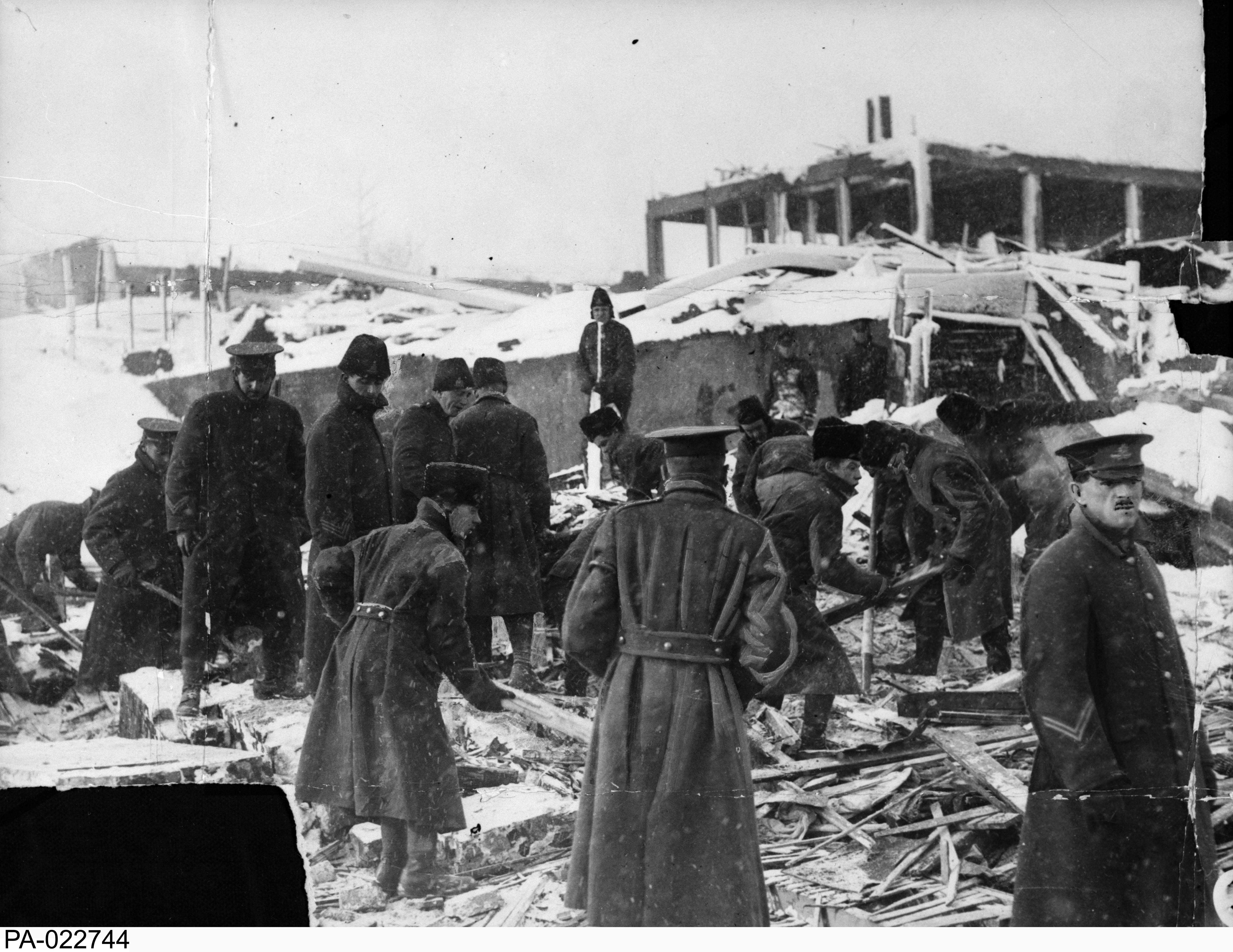 Black and white photograph. A group of soldiers digs through debris, searching for victims of the explosion. They wear long winter coats. The skeletal remains of a building  are visible on the hill beyond them.