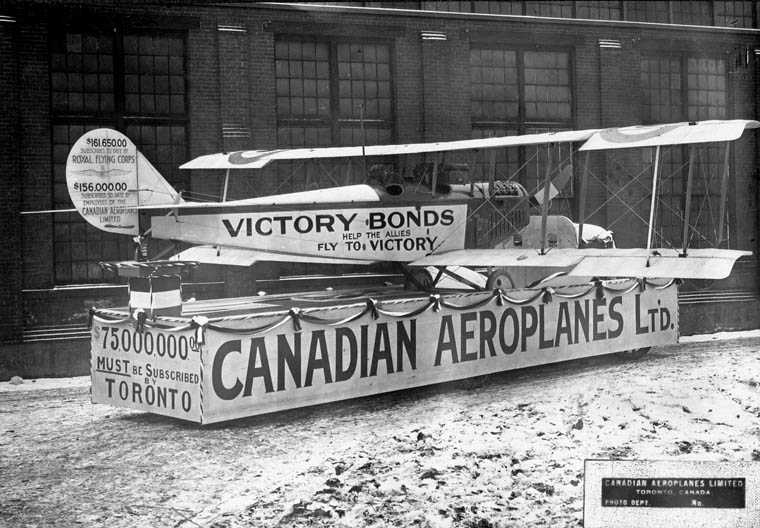 A Canadian airplane sits on a float as part of a Victory Loan parade. Various slogans and information have been printed on it.